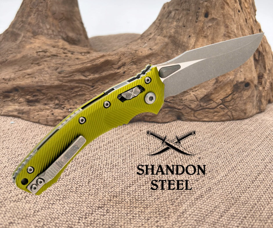 Folding knife with yellow handle and silver blade on a textured surface with 'Shandon Steel' branding.
