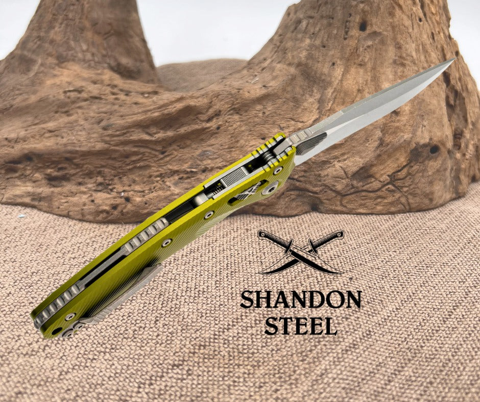 Folding knife with yellow handle and metallic blade on a textured surface, Primemartshop logo visible.