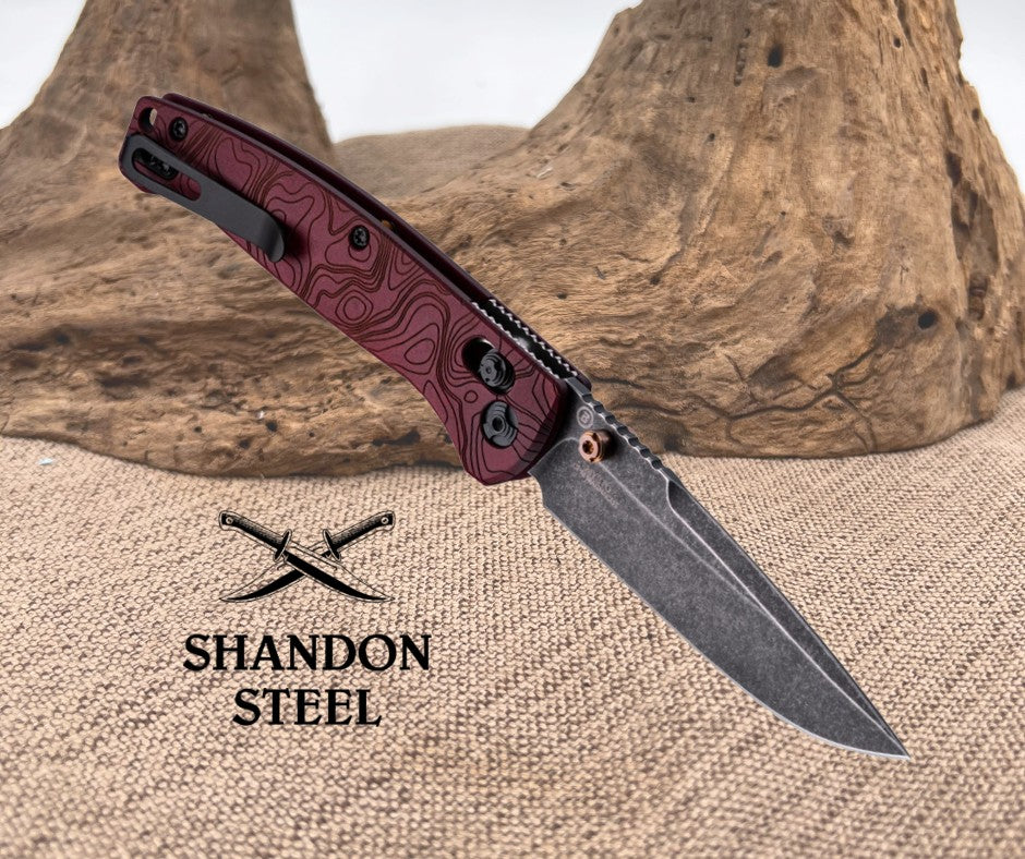 Folding knife with maroon handle and black blade on a textured surface, Primemartshop logo visible.